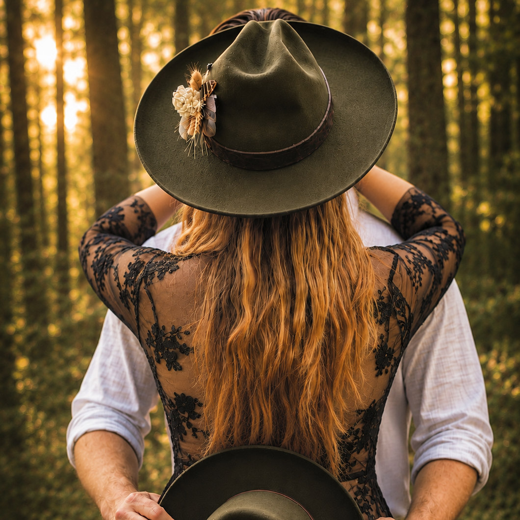 A couple photographed from behind in a sunlit forest, with the woman wearing a wide-brim olive green hat adorned with dried flowers and a black lace backless top over a white shirt. The man wears a white shirt, and both hold a dark hat.