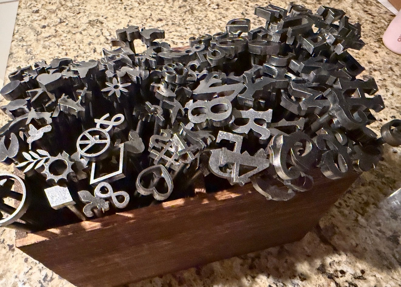 A wooden box filled with many small metal letter and symbol cutters or molds, photographed on a speckled granite countertop.