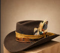 A brown felt cowboy hat with a wide gold and brown decorative band, embellished with feathers, a playing card, and a small embroidered emblem, displayed on a wooden block against a warm beige background.