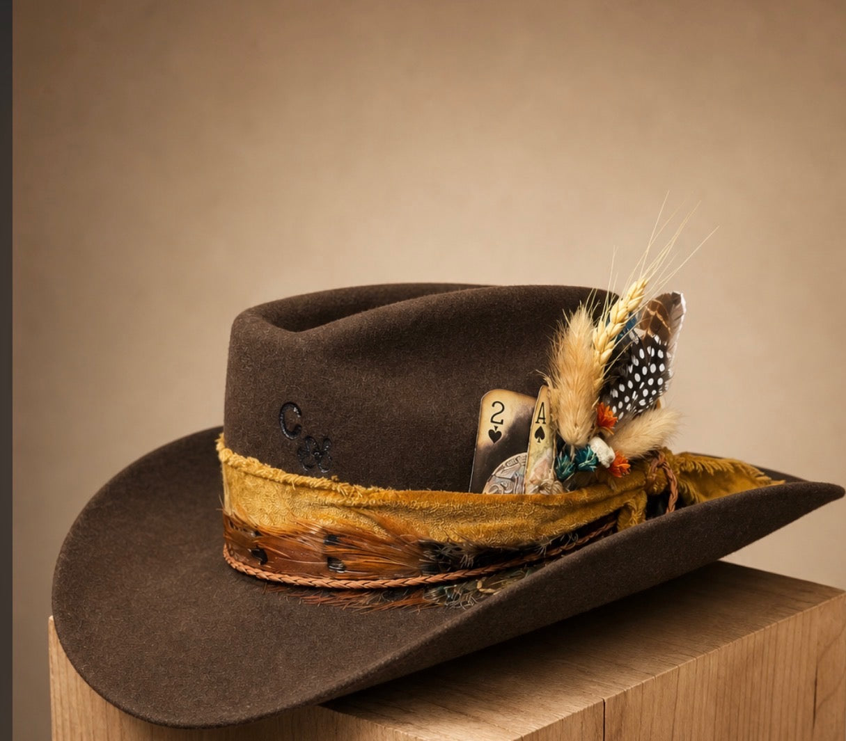 A brown felt cowboy hat with a wide gold and brown decorative band, embellished with feathers, a playing card, and a small embroidered emblem, displayed on a wooden block against a warm beige background.