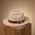 A light beige felt fedora hat with a brown leather band and silver buckle, displayed on a rustic wooden block against a warm beige background.