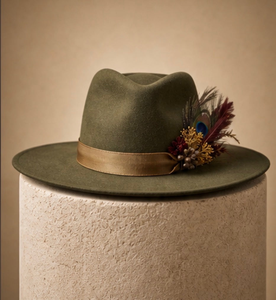 An olive green felt wide-brim hat with a tan ribbon band and a decorative feather and floral accent, displayed on a textured beige cylindrical pedestal against a warm beige background.
