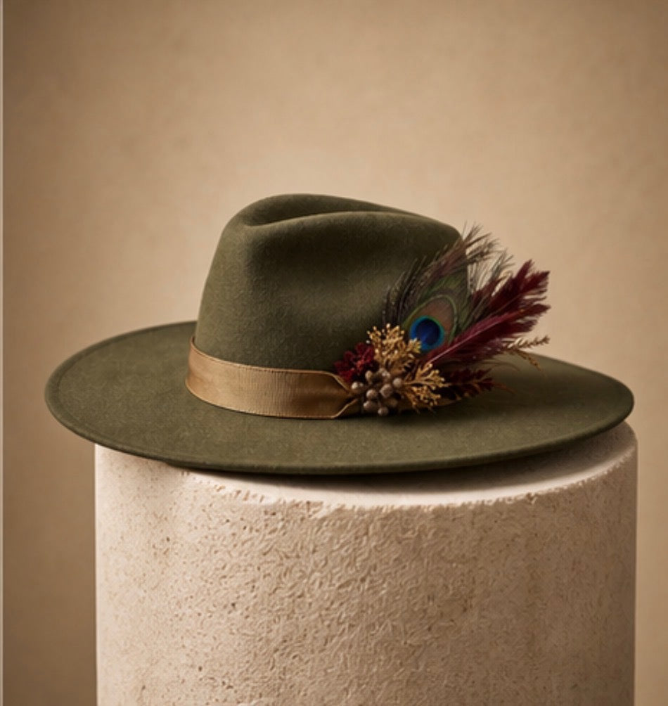 An olive green felt wide-brim hat with a tan ribbon band and a decorative feather and bead cluster, displayed on a textured cream pedestal against a warm beige background.