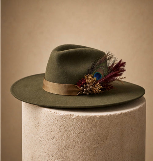 An olive green felt wide-brim hat with a tan ribbon band and a decorative feather and bead cluster, displayed on a textured cream pedestal against a warm beige background.