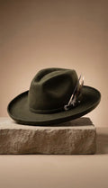 An olive green felt wide-brim hat with a decorative feather accent, resting on a textured stone block against a warm beige background.