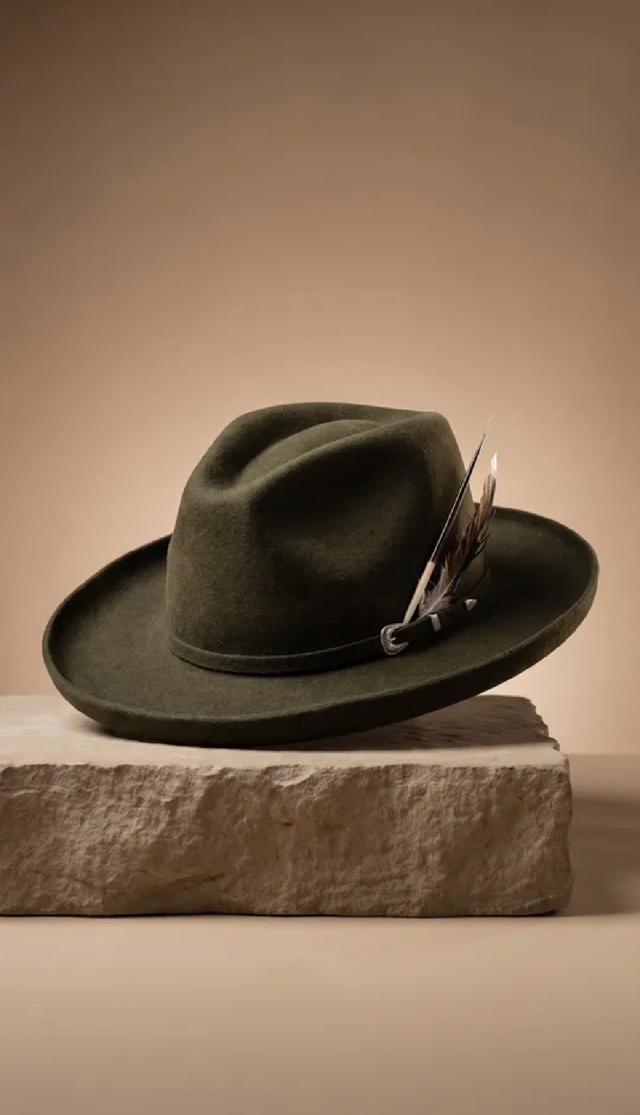 An olive green felt wide-brim hat with a decorative feather accent, resting on a textured stone block against a warm beige background.