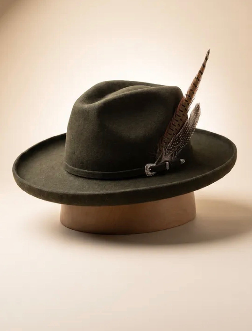 A dark olive green felt wide-brim hat with a decorative brown feather and small silver buckle, displayed on a tan stand against a warm beige background.