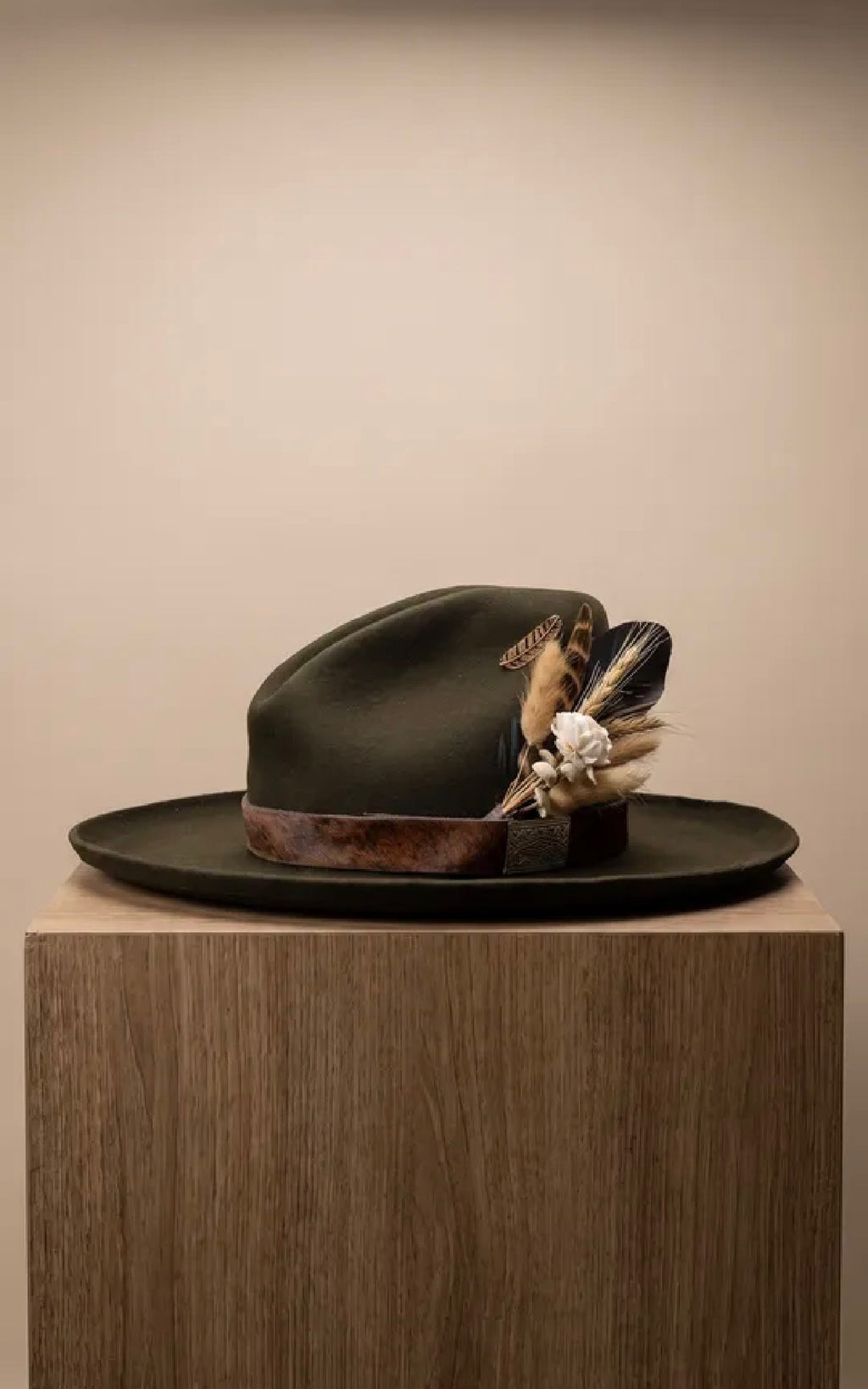 A dark olive green wide-brimmed hat with a brown band and feathered floral embellishments, displayed on a wooden pedestal against a plain beige background.
