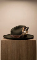 A dark olive green wide-brimmed hat with a brown band and feathered floral embellishments, displayed on a wooden pedestal against a plain beige background.