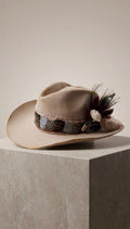 A beige wide-brim fedora hat with a decorative band and feather-and-floral embellishment, displayed on a light stone pedestal against a warm neutral background.