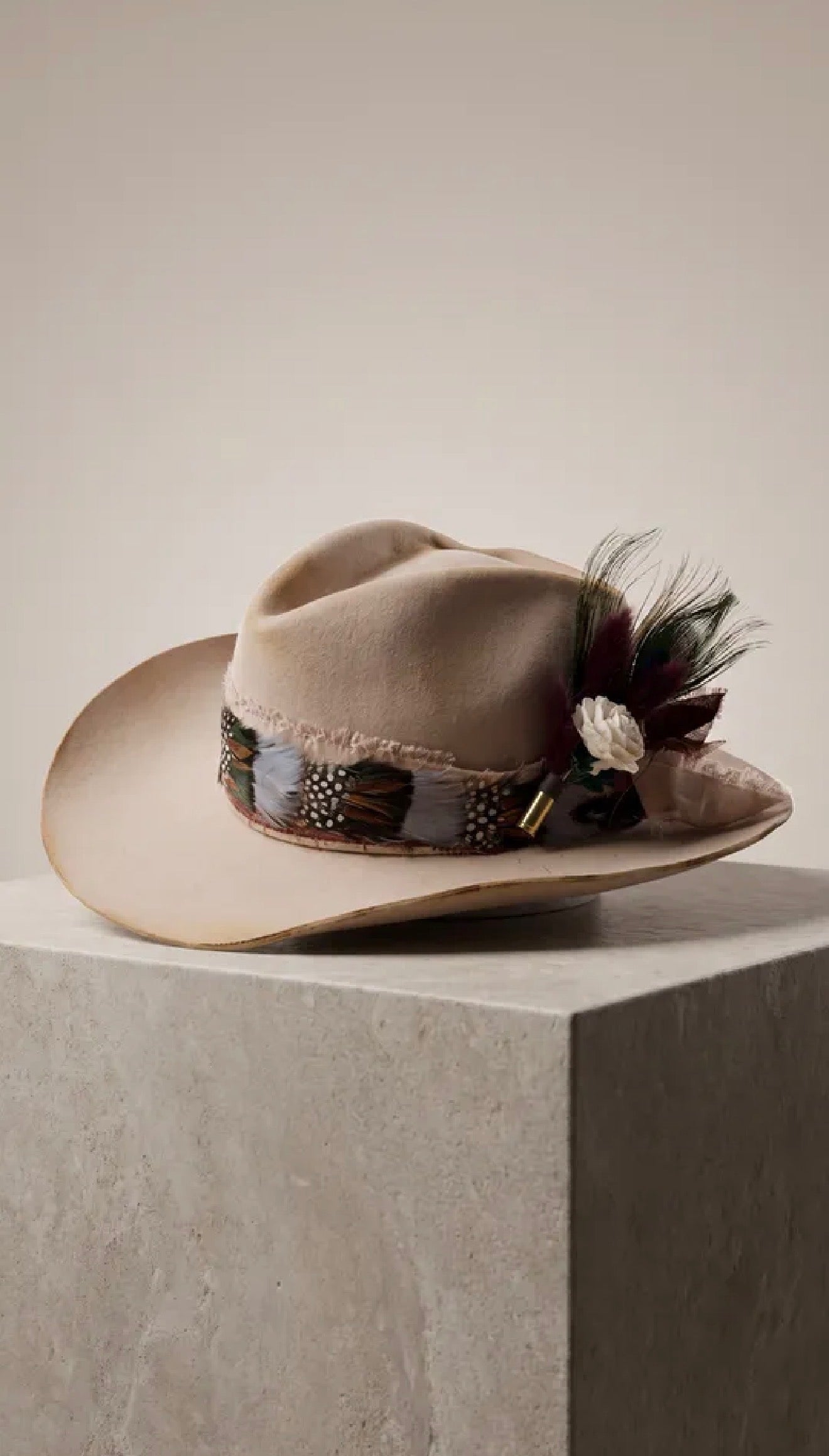 A beige wide-brim fedora hat with a decorative band and feather-and-floral embellishment, displayed on a light stone pedestal against a warm neutral background.