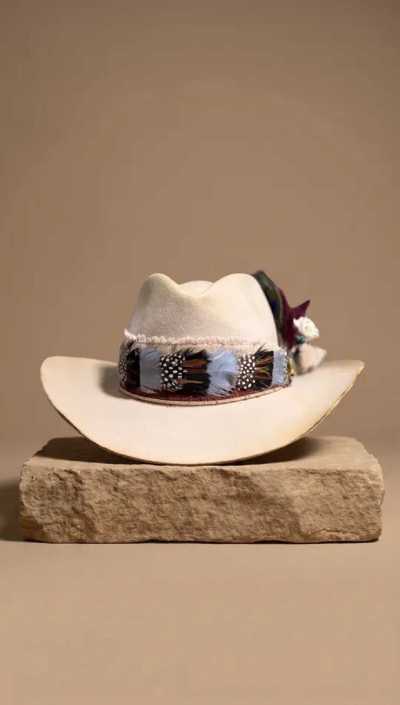 A cream-colored cowboy hat with a decorative band and feather accent, displayed on a rustic stone block against a solid beige background.